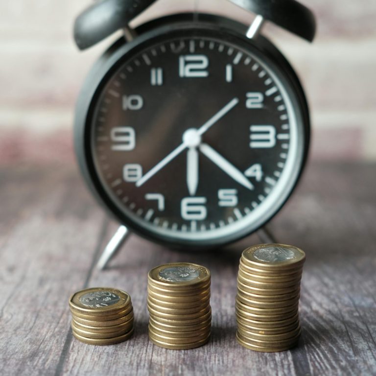 Eine analoge Uhr neben gestapelten Münzen auf einem Holztisch.