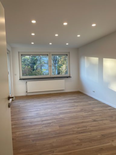 Wohnzimmer Heller, leerer Raum mit großen Fenstern und Holzboden, warmes Licht von Deckenleuchten.